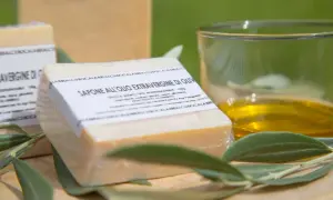 Sapone all'olio extravergine di oliva, artigianale e biodegradabile, accanto a un bicchiere d'olio e foglie d'ulivo.