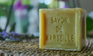 Un pezzo di sapone bucato, un classico sapone di Marsiglia, su una trama di paglia. Sullo sfondo, lavanda e fiori sfocati.