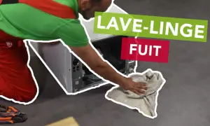 Un tecnico con tuta rossa e verde asciuga con uno straccio il pavimento dove la lavatrice perde acqua dal cestello.