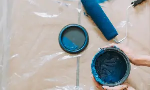 Mani sporche di blu tengono un barattolo di vernice. Un rullo e il coperchio del barattolo, con una macchia di pittura, sono appoggiati su un telo protettivo.