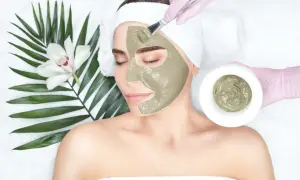 Una donna riceve un trattamento viso con maschera all'argilla verde, applicata con un pennello. Accanto, una foglia di palma e un fiore bianco.
