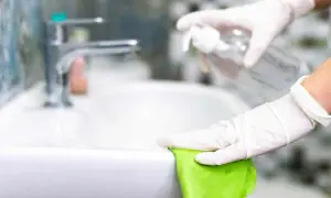 Mani guantate puliscono il lavandino con un panno verde e uno spray, mostrando come pulire il bagno in modo naturale.