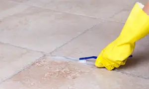 Mano con guanto giallo pulisce le fughe dei pavimenti con uno spazzolino e schiuma.