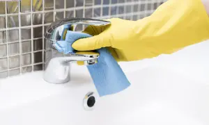 Mano con guanto giallo pulisce rubinetto cromato con panno blu. Come togliere le pietre di sole dal bagno.