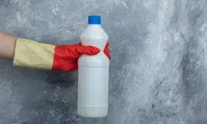 Mano con guanto rosso e giallo tiene una bottiglia bianca di candeggina, pronta per pulire il cemento.