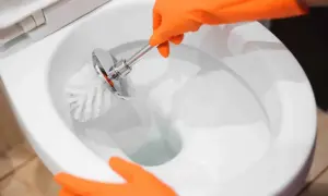 Mani con guanti arancioni puliscono il water, rimuovendo le incrostazioni d'acqua con uno scopino.