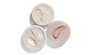 Tre barattoli aperti con texture cremose: un bianco avorio, un grigio chiaro e un rosa perlato. Ideali per un burro corpo nutriente.