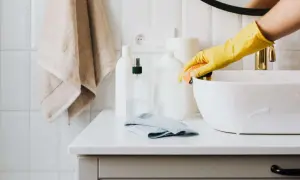 Mano con guanto giallo pulisce lavandino con spugna. Sul piano, flaconi di detersivi fatti in casa e asciugamano.
