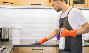 Uomo con guanti arancioni e grembiule pulisce il piano della cucina con uno spray. Ecco come pulire la cucina in modo efficace.