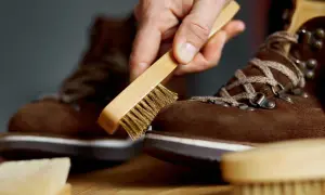 Mano che spazzola una scarpa scamosciata marrone con una spazzola metallica, mostrando come pulire la pelle scamosciata delle scarpe per mantenerla in forma.