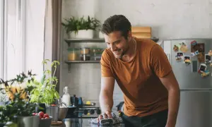 Uomo sorridente pulisce il piano cottura con un panno, mentre sul bancone ci sono piante aromatiche e pomodorini. Forse usa cristalli di soda per lucidare.