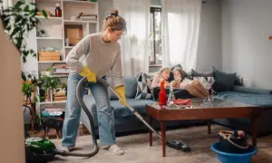 Donna con guanti gialli aspira il tappeto, mentre due bambine guardano il telefono sul divano. Una guida pratica su come pulire una casa sporchissima.