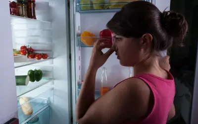 Donna con mano sul naso davanti a un frigorifero pieno di cibo, preoccupata per come togliere la puzza dal frigo.