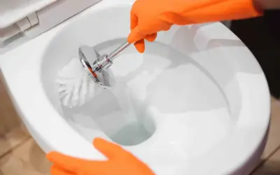 Mani con guanti arancioni puliscono il water, rimuovendo le incrostazioni d'acqua con uno scopino.