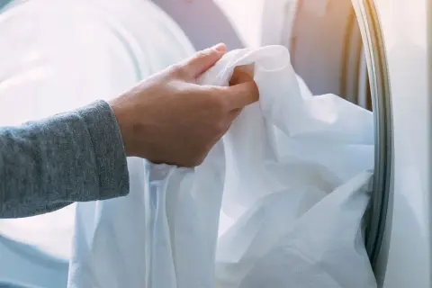 Mano che inserisce un lenzuolo bianco nella lavatrice. Ricorda che candeggina e bicarbonato si possono mischiare con cautela per un bucato più bianco.