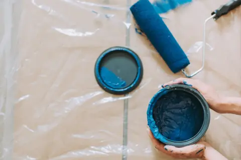 Mani sporche di blu tengono un barattolo di vernice. Un rullo e il coperchio del barattolo, con una macchia di pittura, sono appoggiati su un telo protettivo.