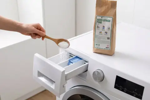 Mano versa un cucchiaio di acido citrico puro nel cassetto della lavastoviglie per una pulizia profonda.