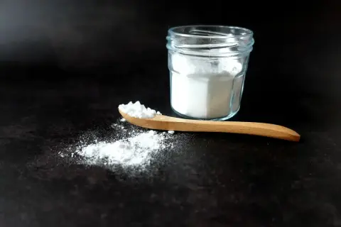 Un cucchiaio di legno con bicarbonato, pronto per smacchiare con bicarbonato. Un barattolo di vetro pieno di polvere bianca sullo sfondo.
