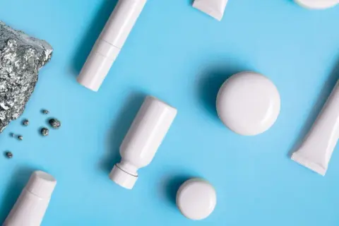 Pietra grezza e piccoli frammenti metallici accanto a flaconi e tubetti bianchi, evocano la purezza dei cosmetici nichel free.