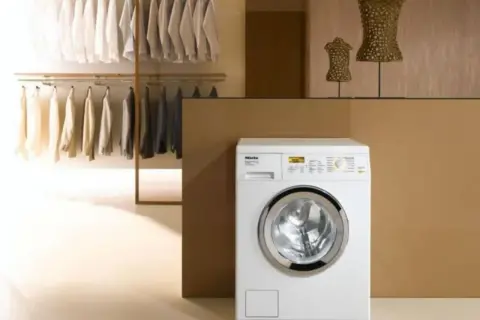 Lavatrice Miele bianca con ingresso acqua calda e fredda, accanto a un armadio con abiti ordinati.