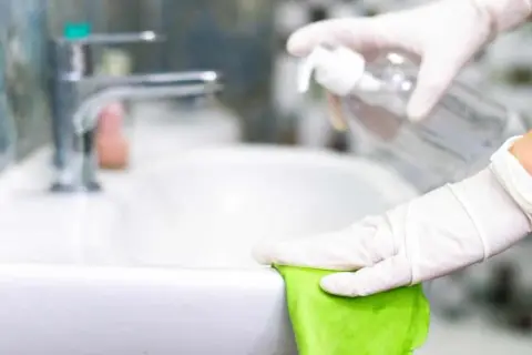 Mani guantate puliscono il lavandino con un panno verde e uno spray, mostrando come pulire il bagno in modo naturale.