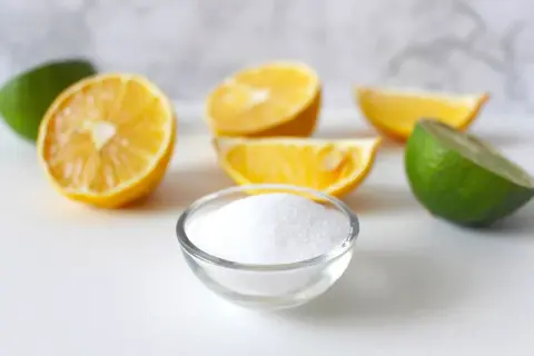 Una ciotola di cristalli bianchi, che ricordano la struttura dell'acido citrico, circondata da spicchi di limone e lime freschi.