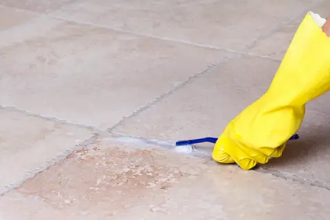 Mano con guanto giallo pulisce le fughe dei pavimenti con uno spazzolino e schiuma.