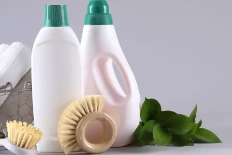 Flaconi bianchi con tappi verdi, spazzole in legno e foglie verdi, per un'idea di detersivi sostenibili.