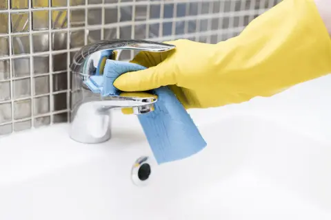 Mano con guanto giallo pulisce rubinetto cromato con panno blu. Come togliere le pietre di sole dal bagno.