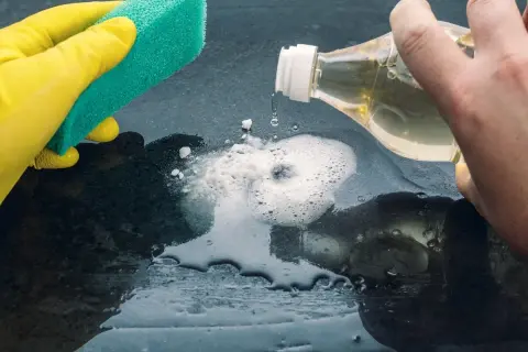 Mano con guanto giallo pulisce con spugna. Aceto sgrassa la superficie, creando schiuma.