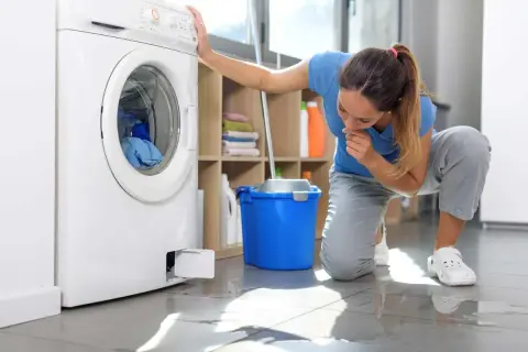 Donna preoccupata guarda la lavatrice che perde acqua dalla vaschetta del detersivo. Sul pavimento si vede una pozza d'acqua.
