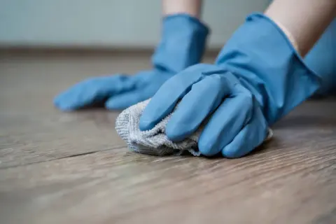 Mani con guanti blu puliscono un pavimento in legno con un panno, creando un detersivo pavimenti fai da te.