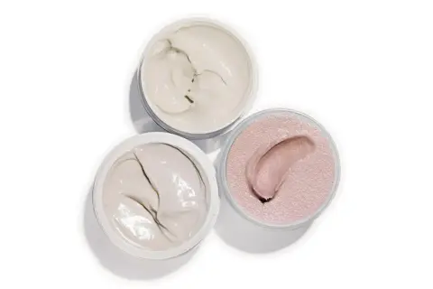 Tre barattoli aperti con texture cremose: un bianco avorio, un grigio chiaro e un rosa perlato. Ideali per un burro corpo nutriente.