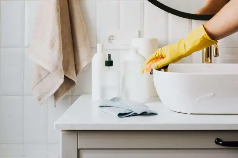 Mano con guanto giallo pulisce lavandino con spugna. Sul piano, flaconi di detersivi fatti in casa e asciugamano.