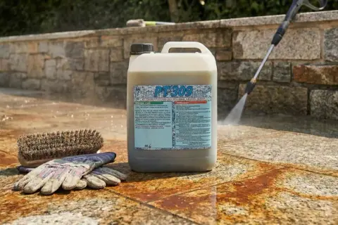 Un flacone di PT309, uno sgrassatore che va risciacquato, è posizionato su un patio in pietra. Accanto, un guanto da lavoro e una spazzola. In sottofondo, un idropulitrice spruzza acqua.