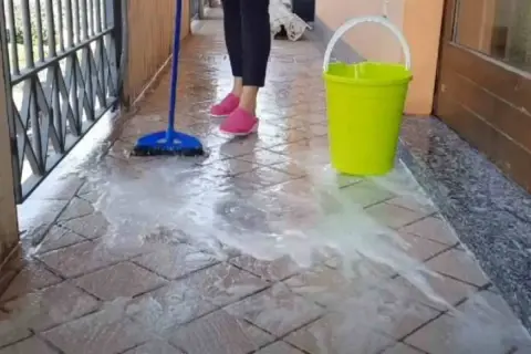 Pulizia del terrazzo esterno: una scopa blu lava le piastrelle con acqua saponata, accanto un secchio giallo.