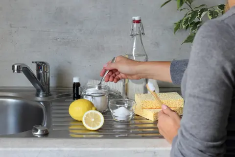 Mano che preleva bicarbonato da un barattolo, con limone, percarbonato e spugne. Differenza tra percarbonato e bicarbonato per pulizie ecologiche.