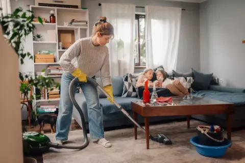 Donna con guanti gialli aspira il tappeto, mentre due bambine guardano il telefono sul divano. Una guida pratica su come pulire una casa sporchissima.