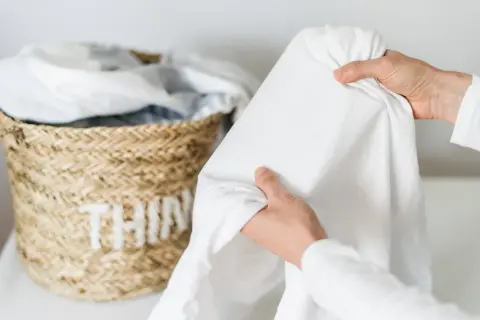 Mani che piegano un lenzuolo bianco, vicino a un cesto di vimini con biancheria. Speriamo non ci siano macchie candeggina!