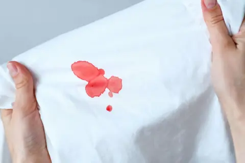 Mani che tengono un tessuto bianco con macchie di sangue rosso vivo. Scopri come togliere le macchie di sangue in modo efficace.