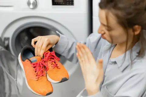 Donna con espressione disgustata tiene in mano scarpe da ginnastica arancioni, pronte per essere lavate in lavatrice.