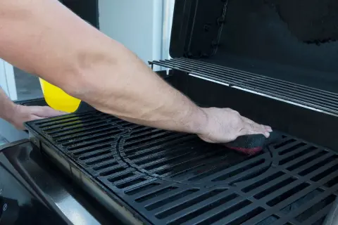 Uomo pulisce la griglia della barbecue con una spugna. Un liquido giallo viene versato da un contenitore.