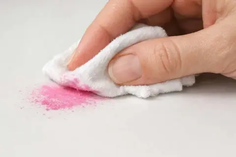 Mano che pulisce polvere rosa da superficie bianca con panno. Scopri come togliere lo smalto dal pavimento in gres.