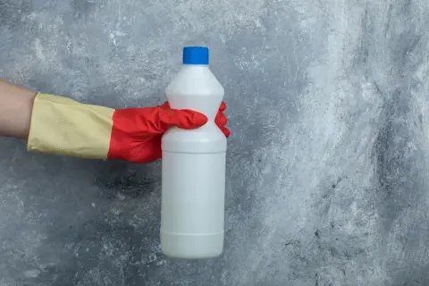 Mano con guanto rosso e giallo tiene una bottiglia di candeggina, pronta per la lavatrice.