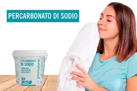 Donna annusa un asciugamano bianco fresco, accanto a un contenitore di percarbonato di sodio. Il percarbonato si può usare sui colorati per un bucato impeccabile.