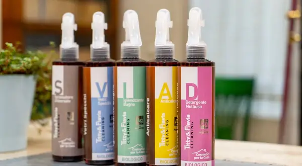 Cinque flaconi spray con etichette colorate: "S" (Formulatore Universale), "V" (Vetri Specchi), "I" (Igienizzante Bagno), "A" (Anticalcare), "D" (Detergente Multiuso). La differenza tra detergente e detersivo è chiara: qui sono tutti detergenti biologici.