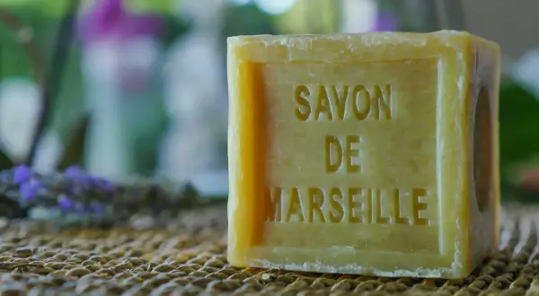 Un pezzo di sapone bucato, un classico sapone di Marsiglia, su una trama di paglia. Sullo sfondo, lavanda e fiori sfocati.