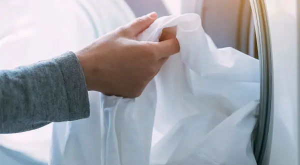 Mano che inserisce un lenzuolo bianco nella lavatrice. Ricorda che candeggina e bicarbonato si possono mischiare con cautela per un bucato più bianco.