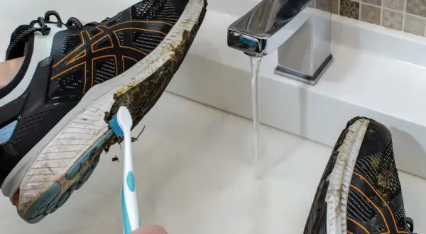 Come lavare le scarpe da running: una mano con uno spazzolino pulisce il fango dalla suola di una scarpa sotto un rubinetto aperto.