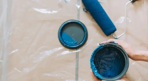 Mani sporche di blu tengono un barattolo di vernice. Un rullo e il coperchio del barattolo, con una macchia di pittura, sono appoggiati su un telo protettivo.